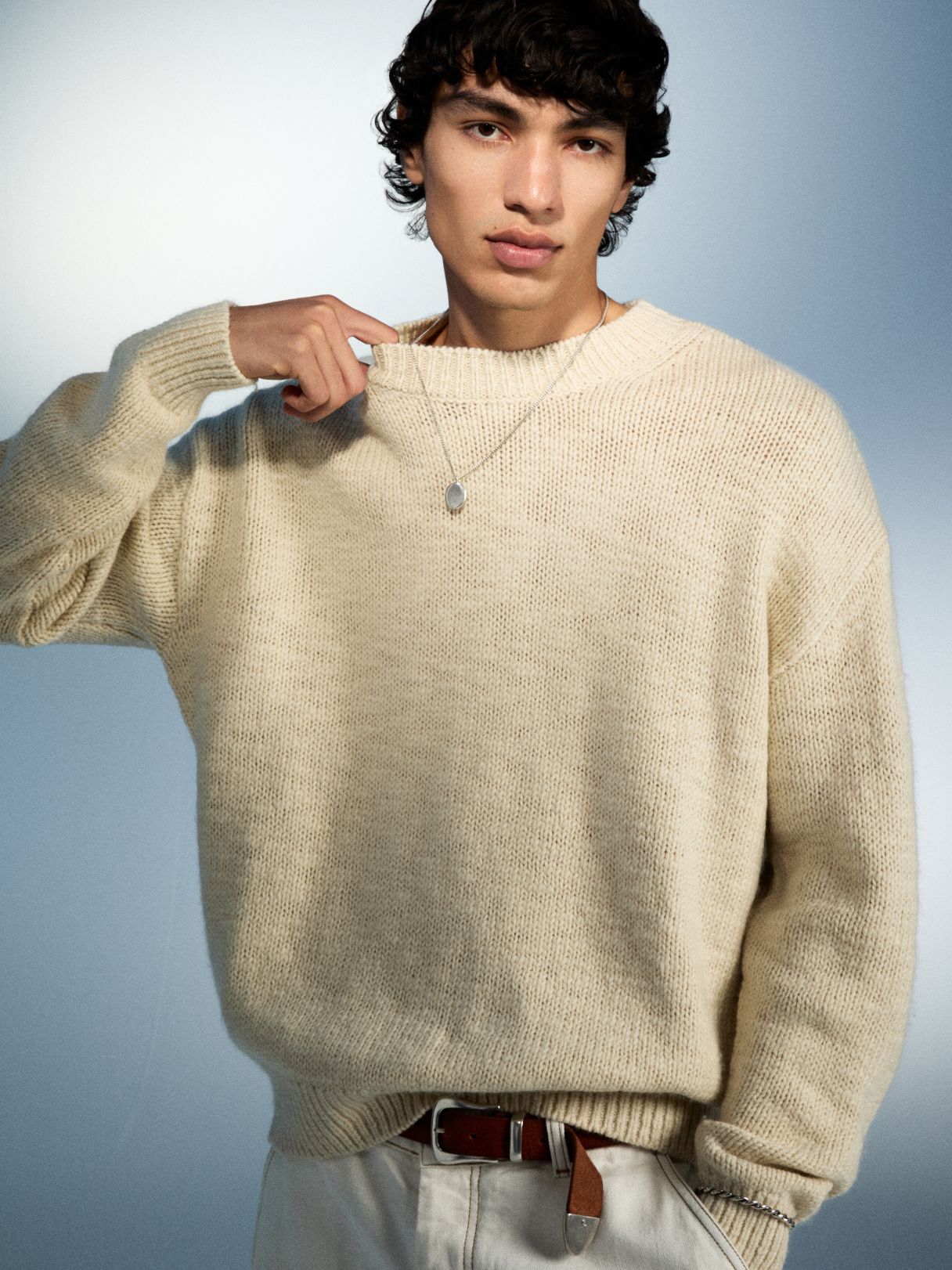 Ein junger Mann mit dunklen Haaren trägt einen cremefarbenen Pullover und eine Halskette.