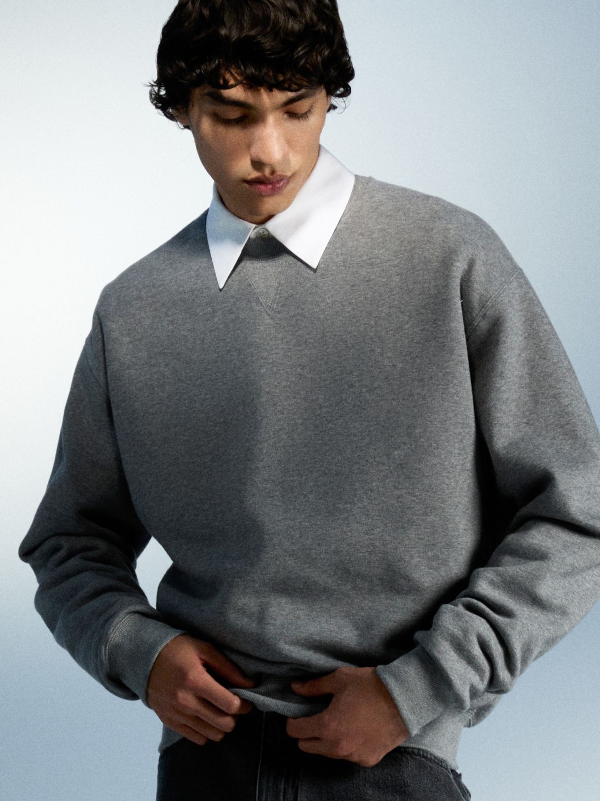 Ein junger Mann mit dunklen Locken trägt einen grauen Pullover über einem weißen Hemdkragen.