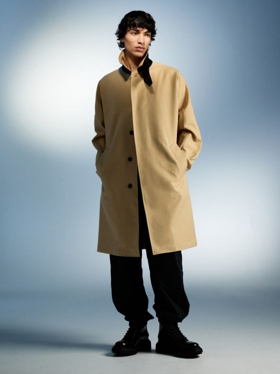 Ein Mann in einem beigen Trenchcoat und schwarzen Hosen steht vor einem blauen Hintergrund.
