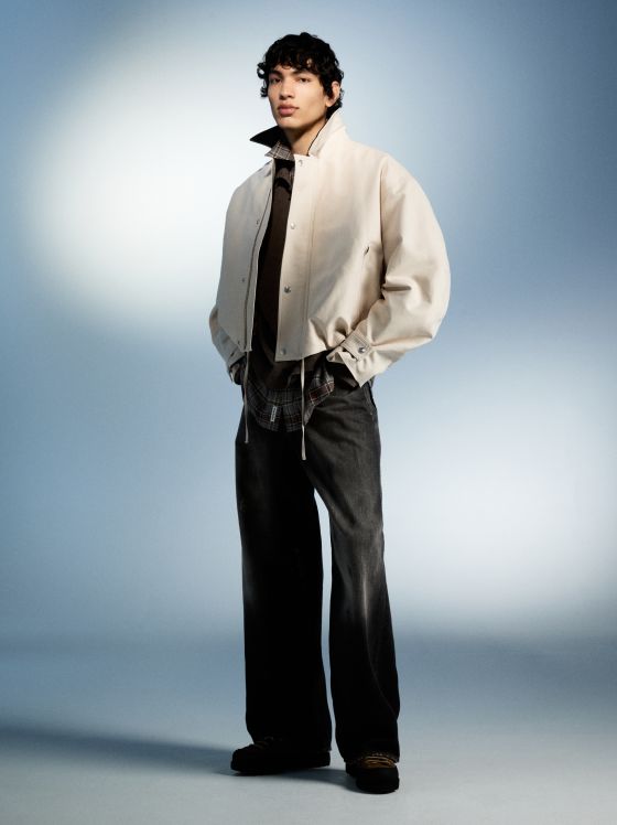 Ein Mann mit lockigem Haar trägt eine cremefarbene Jacke, ein braunes Hemd und weite Jeans.