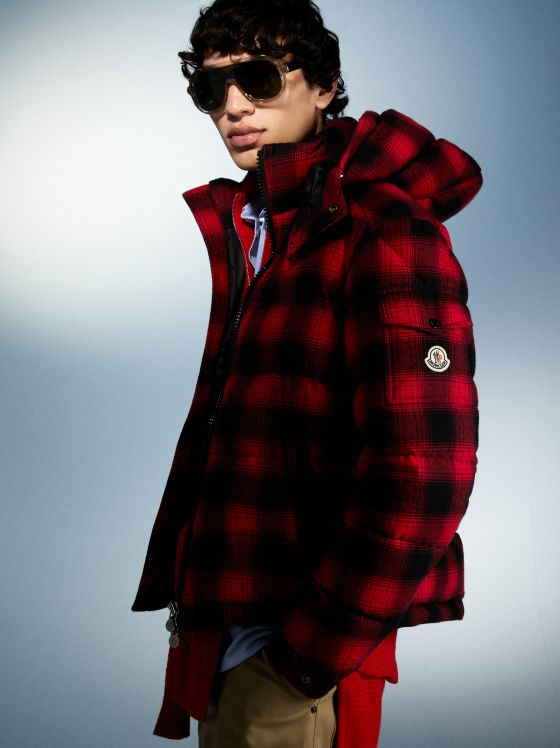 Mann mit karierter Moncler-Jacke und Sonnenbrille, stehend vor blauem Hintergrund.