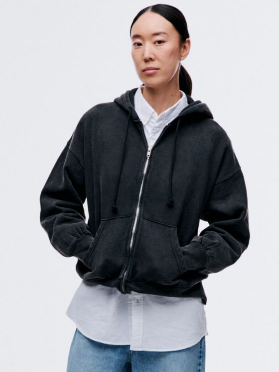 Eine Frau mit Pferdeschwanz trägt einen schwarzen Reißverschluss-Hoodie über einem weißen Hemd und Jeans.