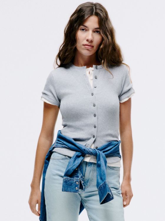 Frau mit braunen Haaren trägt hellgraues Cardigan über weißem Top und Jeans.